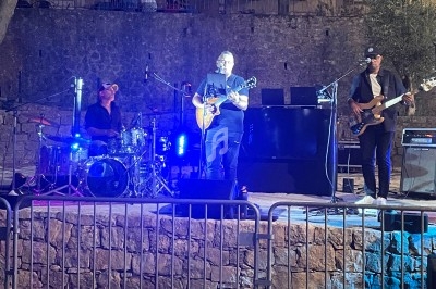 Un groupe de musiciens joue en plein air sur une scène éclairée, avec un guitariste, un batteur et un bassiste.