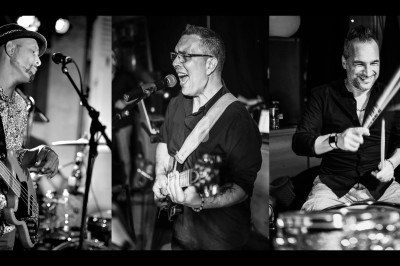 Trois musiciens jouant en concert, avec un bassiste, un chanteur-guitariste et un batteur, en noir et blanc.