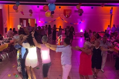 Des personnes dansent en chaîne dans une salle décorée de guirlandes et lanternes colorées, éclairée par des lumières vives.