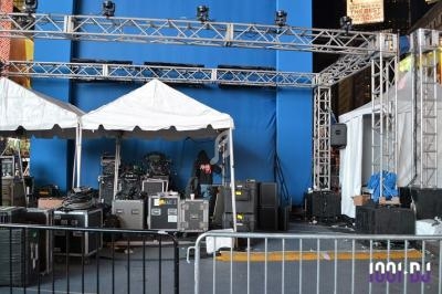 Scène extérieure en préparation avec tentes blanches, matériel audio et éclairage sous une structure métallique.