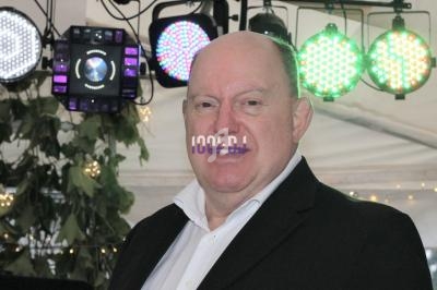 Homme en costume noir devant des lumières colorées et des plantes dans un environnement festif.