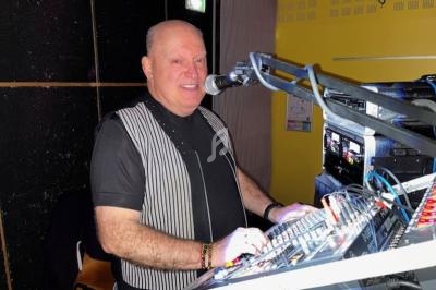 Homme souriant devant une console de mixage, parlant dans un micro, dans un environnement intérieur éclairé.