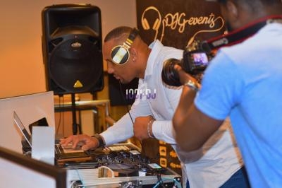 Un DJ mixe de la musique sur une console, portant un casque audio, tandis qu'une personne filme avec une caméra.