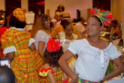 Des femmes et une enfant en tenues traditionnelles colorées dansent et sourient lors d'un événement festif en intérieur.