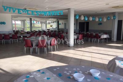 Salle de réception décorée avec des tables alignées, nappes grises, serviettes bleues et chaises rouges.