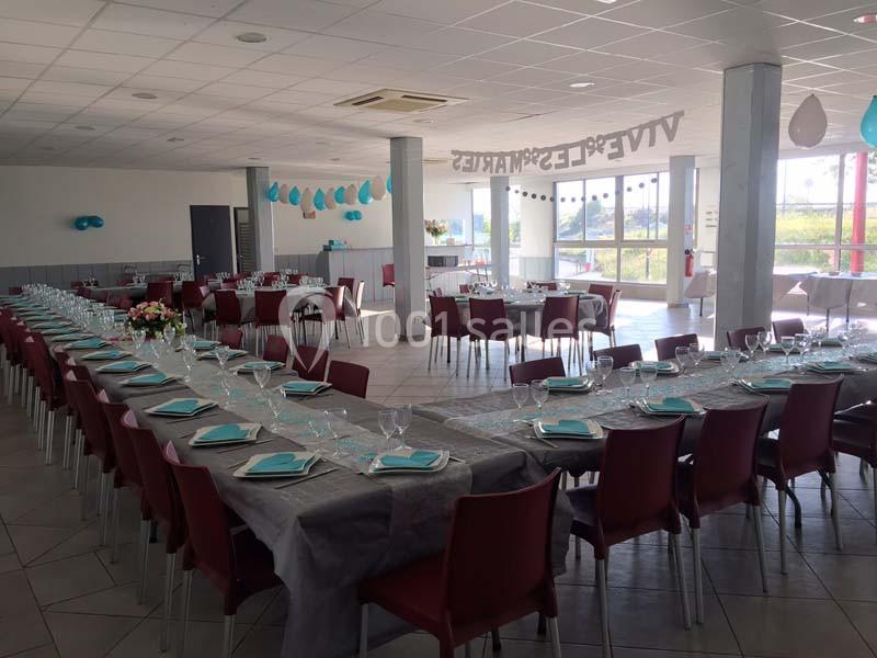 Salle de réception décorée avec des tables alignées, nappes grises, serviettes turquoise et ballons suspendus.