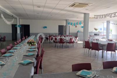 Salle de réception décorée avec des tables alignées, nappes grises, serviettes bleues et chaises rouges.