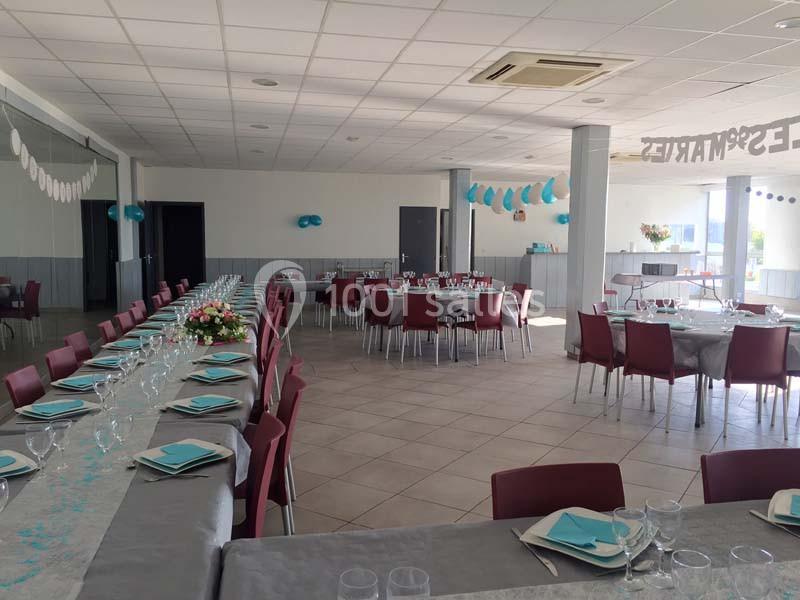 Salle décorée pour un événement avec tables dressées, nappes grises, serviettes bleues et guirlandes suspendues.