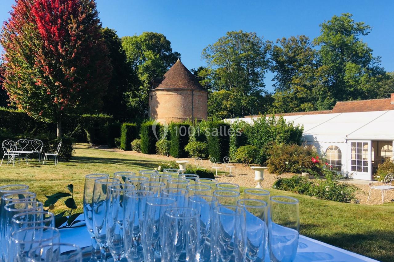 Table avec des verres vides en extérieur, devant un jardin verdoyant, un bâtiment en briques et une tente blanche.