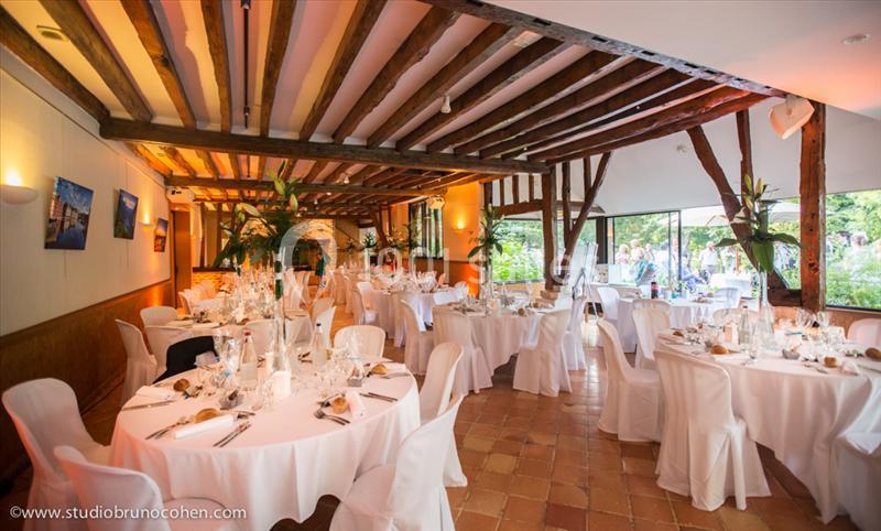 Salle de réception lumineuse avec tables rondes décorées pour un événement, poutres apparentes et grandes baies vitrées.