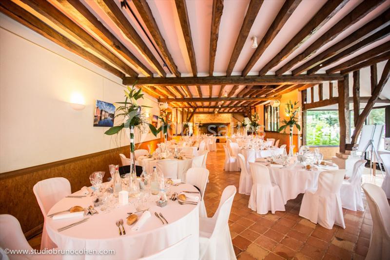 Salle de réception lumineuse avec tables rondes dressées, chaises blanches et poutres apparentes au plafond.