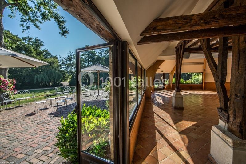 Vue depuis une salle avec poutres apparentes donnant sur une terrasse ensoleillée avec tables, chaises et jardin verdoyant.