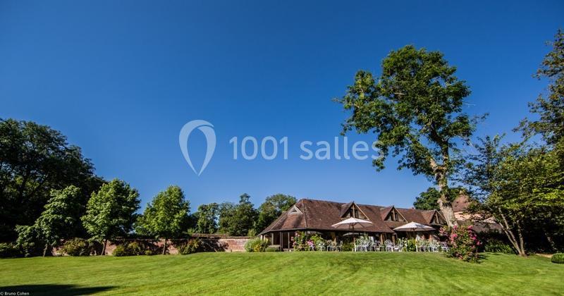 Pelouse verdoyante avec une maison en briques et toit en tuiles, entourée d'arbres et de parasols sur une terrasse.
