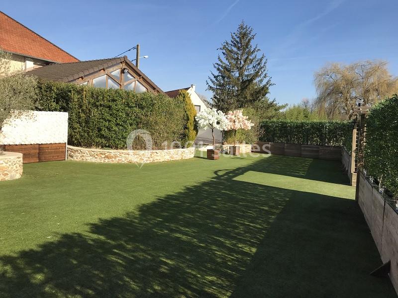 Jardin avec pelouse synthétique, haies taillées, arbres décoratifs et terrasse en arrière-plan sous un ciel dégagé.