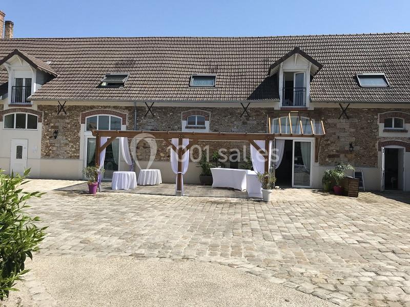 Façade d'une bâtisse en pierre avec toit en tuiles, terrasse pavée et pergola décorée de voilages blancs.