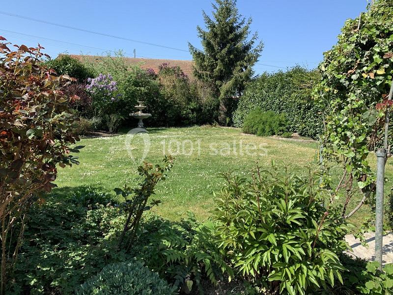 Jardin verdoyant avec pelouse, arbustes, haies, un arbre au fond et une fontaine en pierre sous un ciel dégagé.