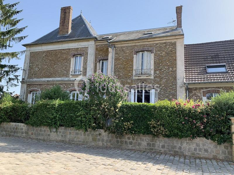 Façade d'une maison en pierre avec volets blancs, entourée de haies fleuries et située près d'une rue pavée.