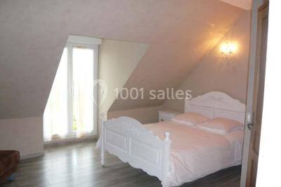 Chambre mansardée avec un lit double blanc, parquet au sol, mur beige et lumière naturelle provenant d'une porte-fenêtre.