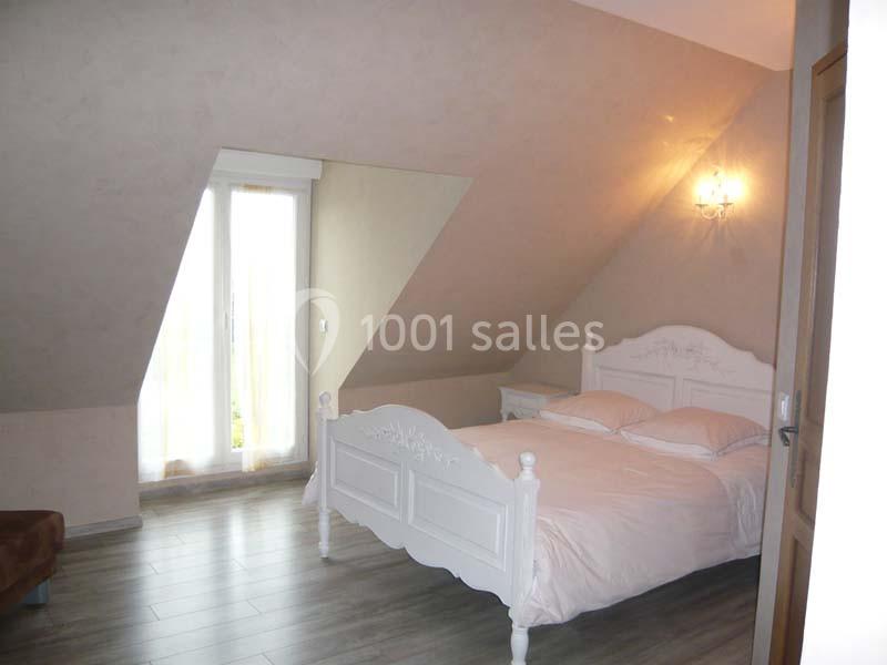 Chambre mansardée avec un lit double blanc, parquet au sol, mur beige et lumière naturelle provenant d'une porte-fenêtre.