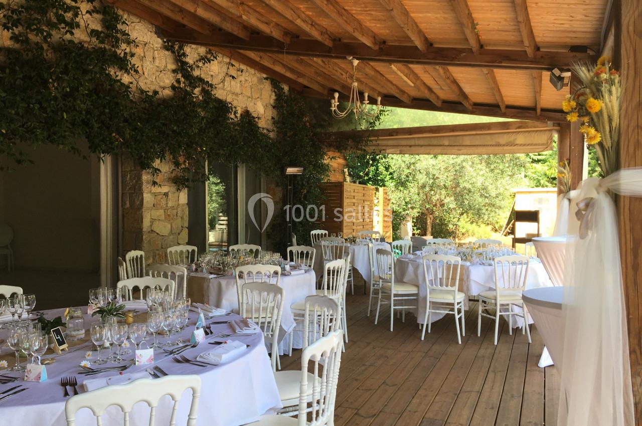 Salle de réception en bois avec tables rondes dressées, chaises blanches et décoration florale, sous une pergola ouverte.