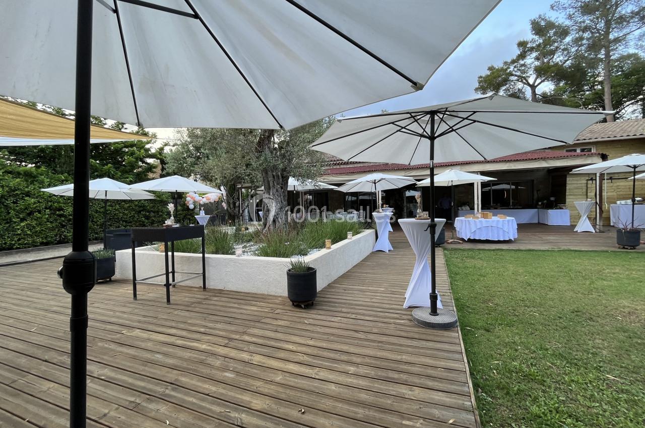 Terrasse en bois avec parasols blancs, espace vert et tables dressées pour un événement en extérieur.