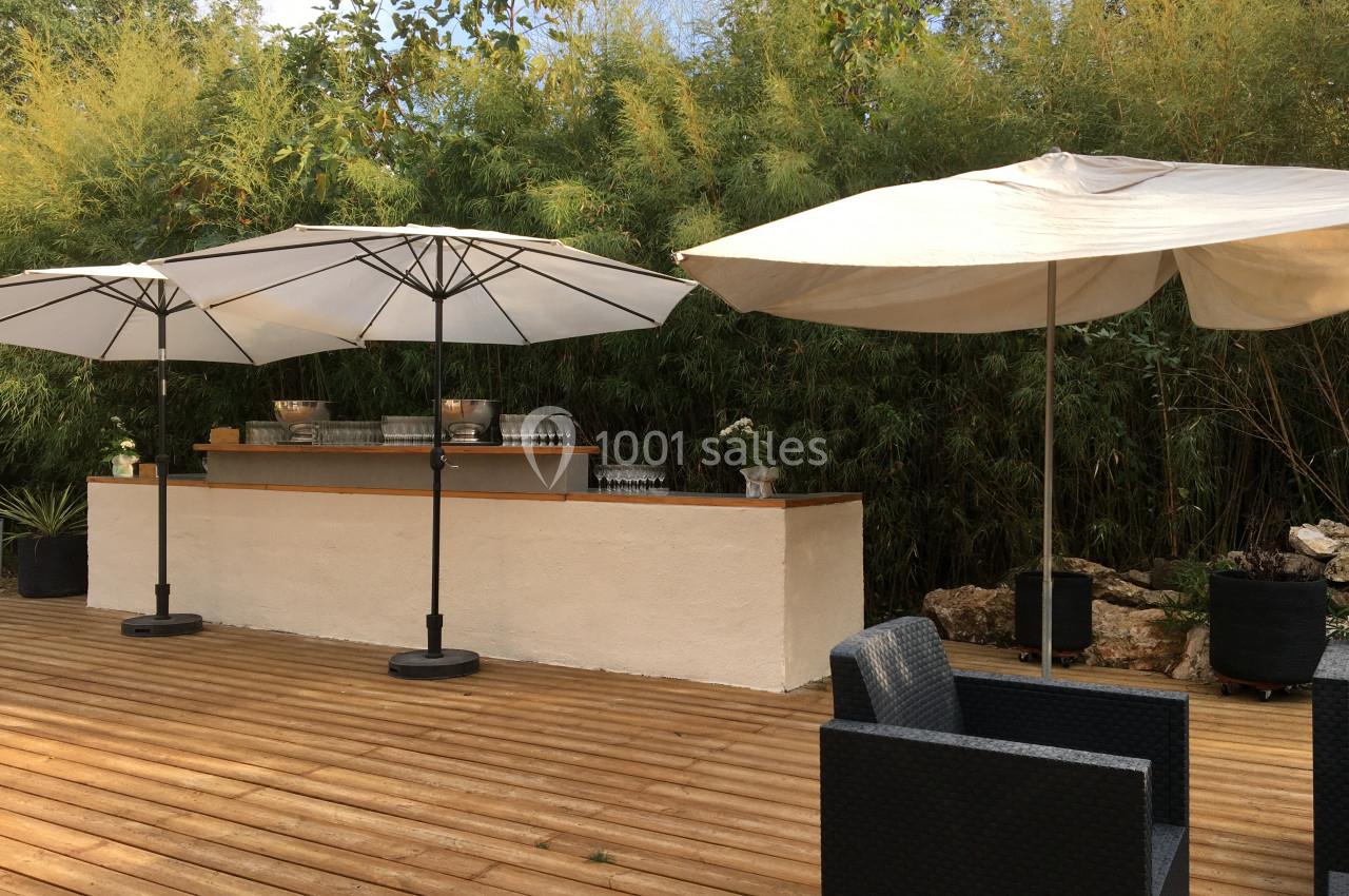 Terrasse en bois avec un bar extérieur, des parasols blancs et des fauteuils noirs, entourée de végétation dense.