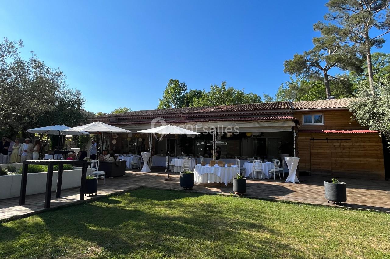 Terrasse en bois d'un bâtiment entouré de végétation, avec tables dressées et parasols sous un ciel bleu.
