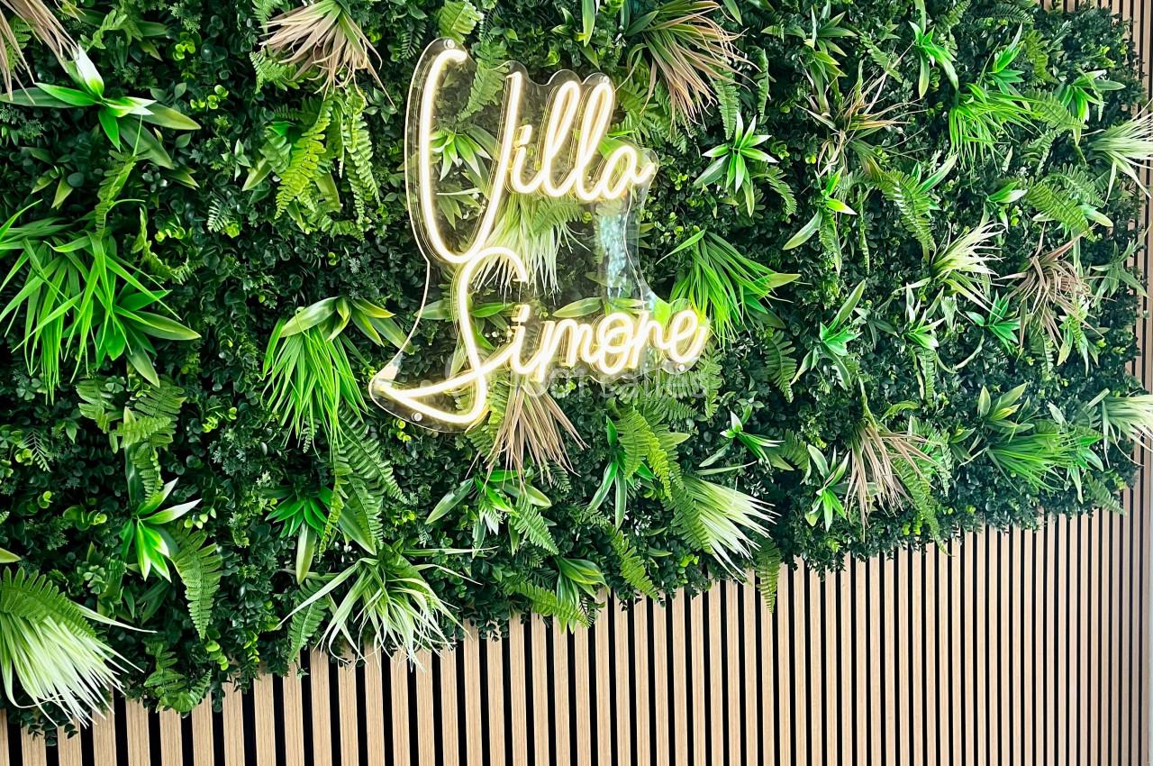 Mur végétal avec des plantes vertes et un néon lumineux indiquant ’Villa Simone’, sur un fond de lattes en bois.