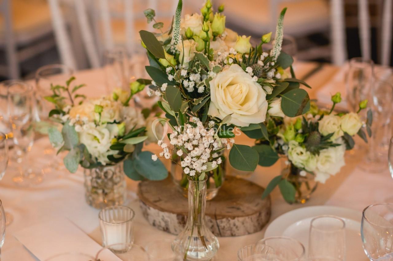 Centre de table floral avec roses blanches, gypsophile et feuillage, entouré de verres et couverts sur une nappe beige.