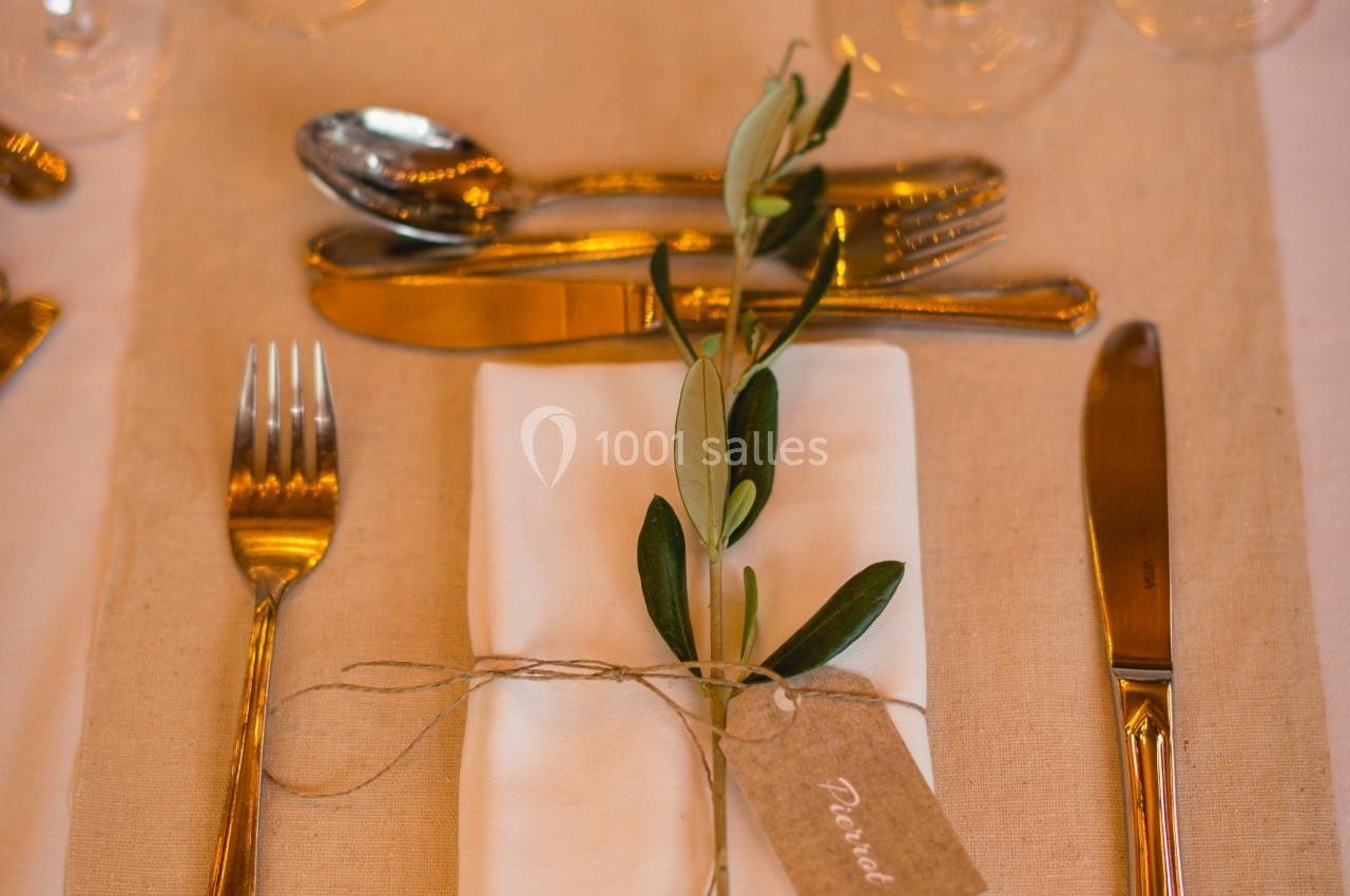 Serviette blanche pliée avec un brin d'olivier et une étiquette, entourée de couverts et verres sur une table dressée.