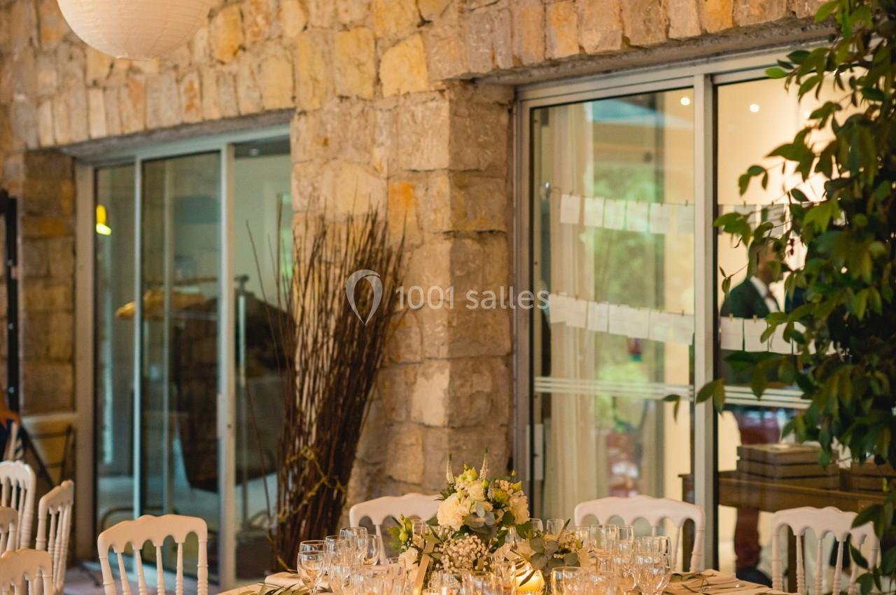Table ronde élégamment dressée avec vaisselle raffinée, décorée de fleurs, dans une salle en pierre avec grandes baies…