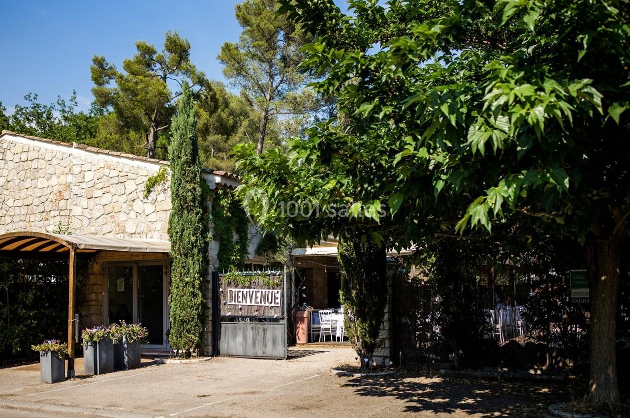 Façade en pierre d'un bâtiment entouré d'arbres, avec une pancarte ’Bienvenue’ et une terrasse ombragée.