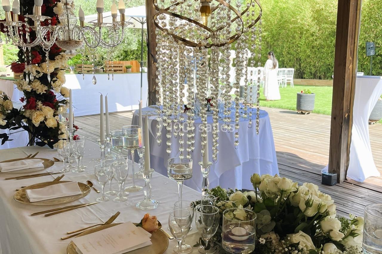 Table élégamment dressée avec vaisselle dorée, chandeliers, fleurs blanches et rouges sous une pergola en bois.