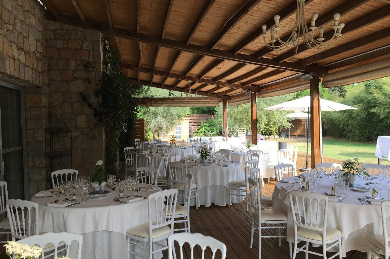 Salle de réception en plein air avec tables rondes dressées, chaises blanches et toit en bois, donnant sur un jardin…