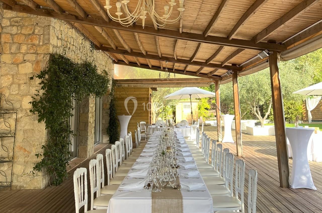 Table longue dressée avec nappes blanches sous une pergola en bois, entourée de verdure et de lumière naturelle.