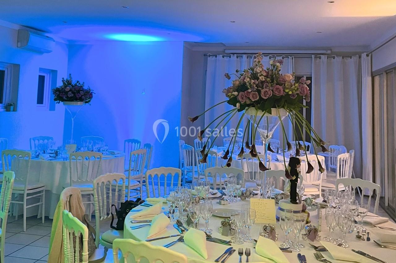 Salle de réception décorée avec des tables dressées, des chaises blanches et des centres de table fleuris.