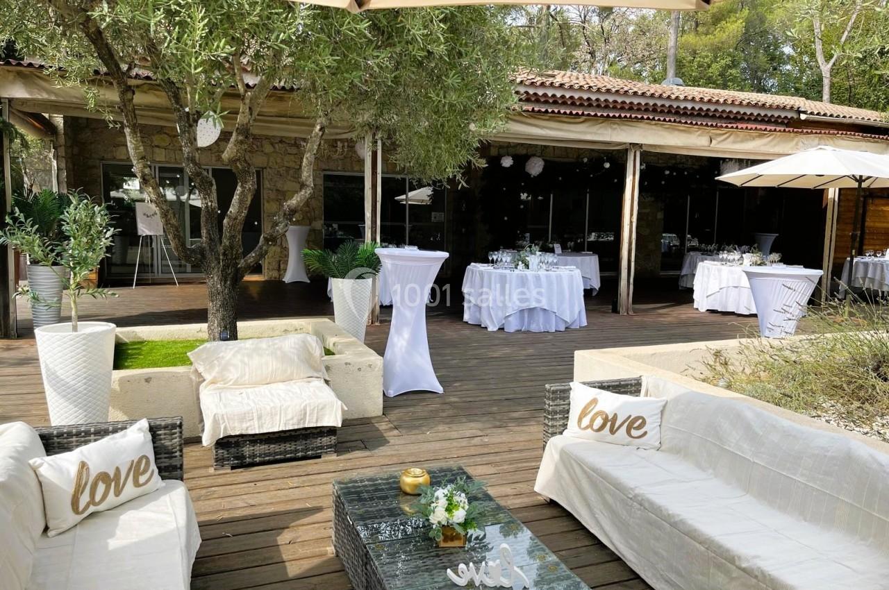 Terrasse en bois aménagée avec canapés, tables hautes et décorations, entourée de végétation et abritée par des parasols.