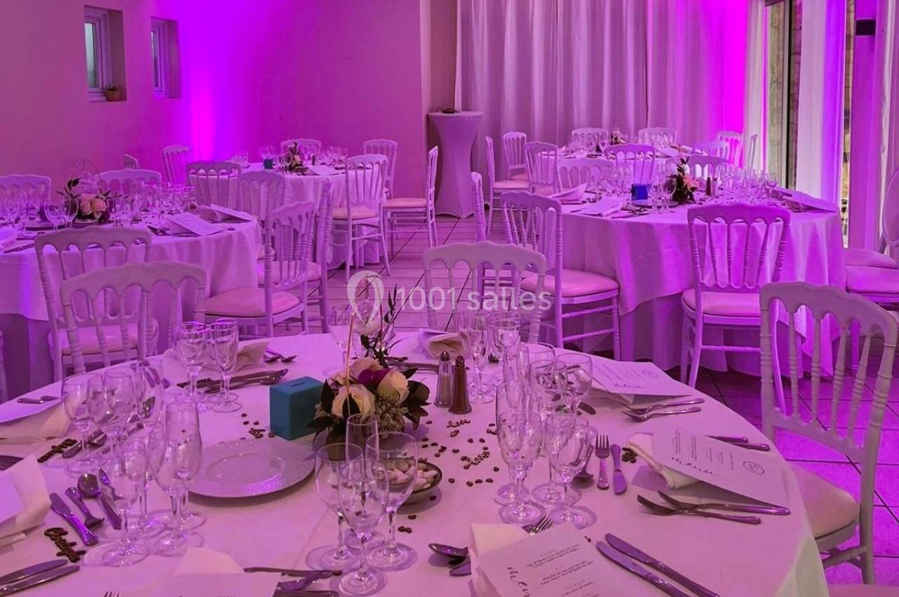 Salle de réception décorée avec des tables rondes dressées, éclairage rose et chaises blanches.