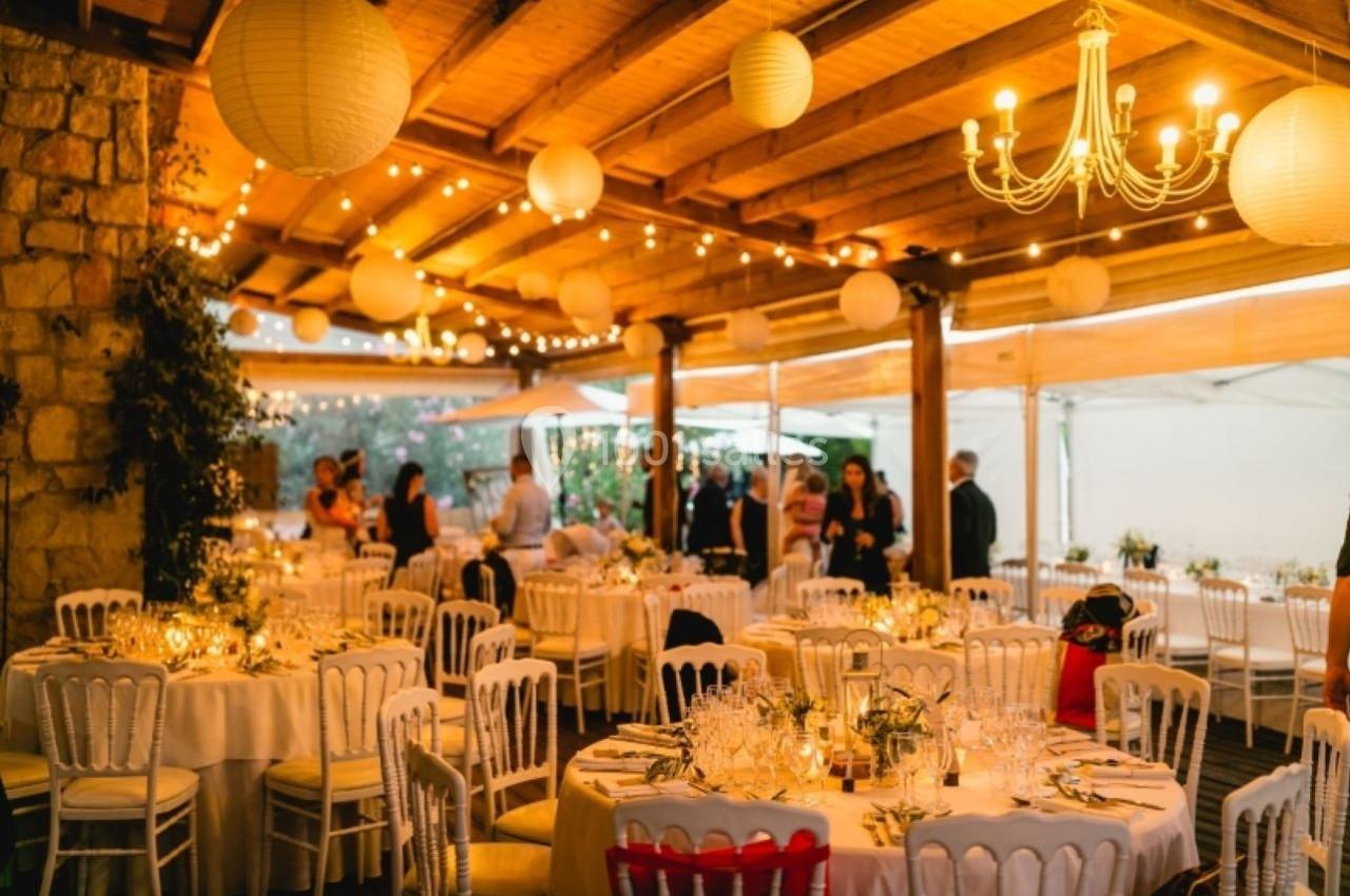 Salle de réception décorée avec des guirlandes lumineuses, des lanternes suspendues et des tables dressées pour un événement.