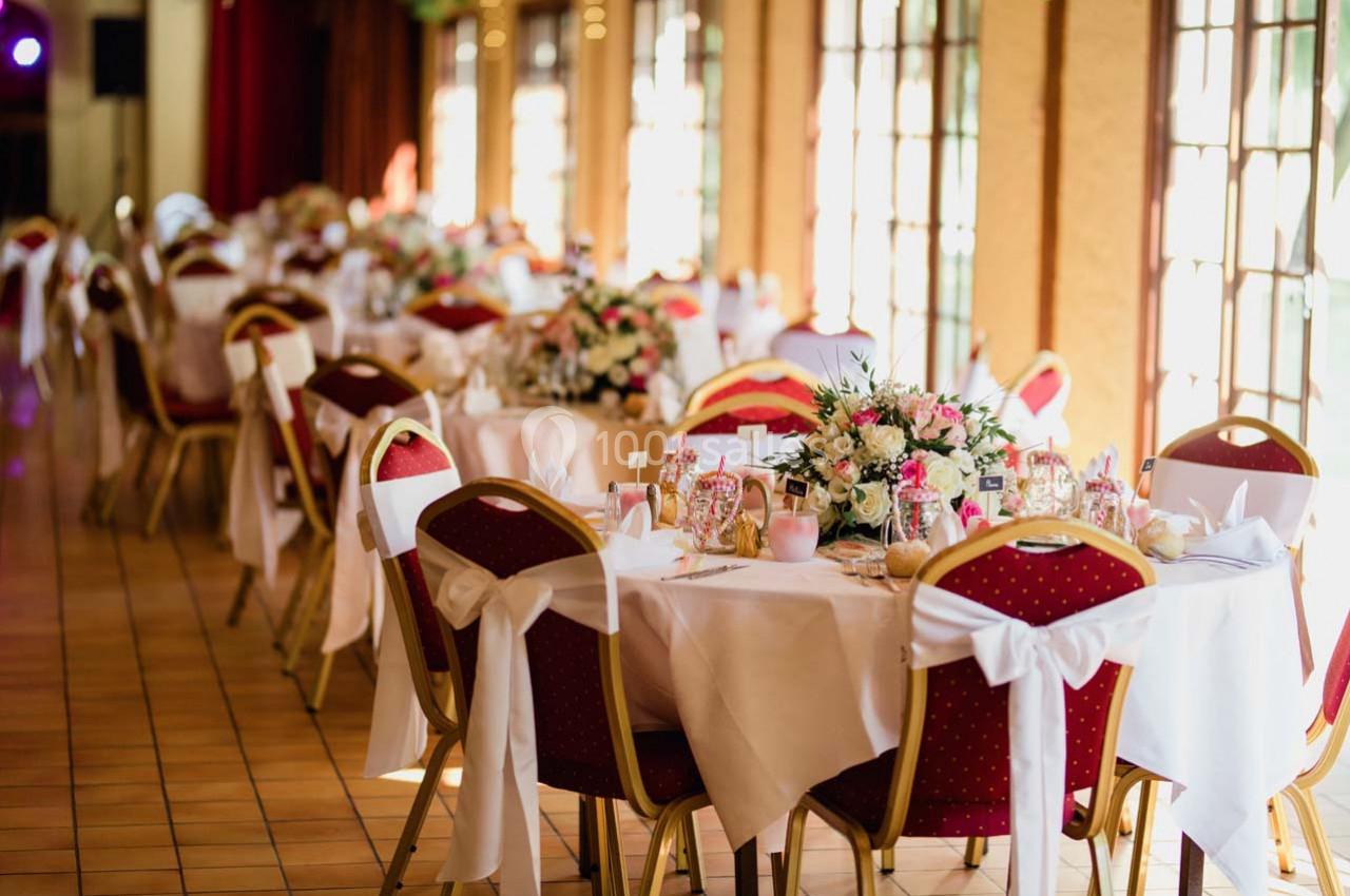 Salle de réception décorée avec des tables rondes, nappes blanches, chaises rouges et centres de table fleuris.