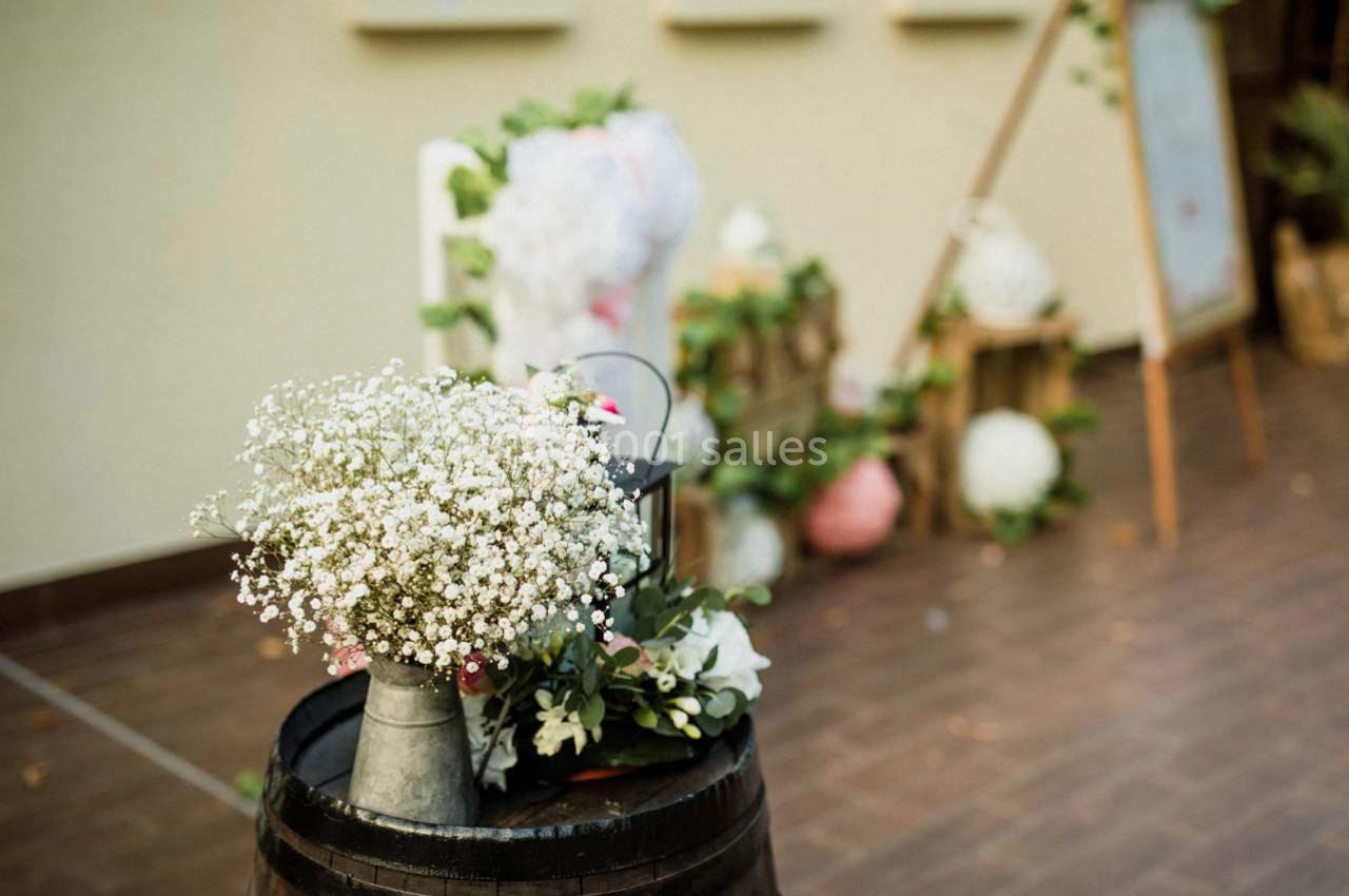 Bouquet de gypsophiles dans un vase en métal posé sur un tonneau, avec une décoration florale floue en arrière-plan.