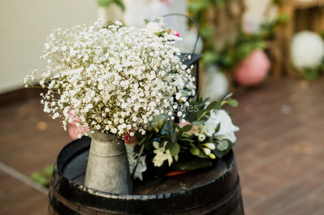 Bouquet de gypsophiles dans un vase métallique posé sur un tonneau en bois, avec des décorations floues en arrière-plan.