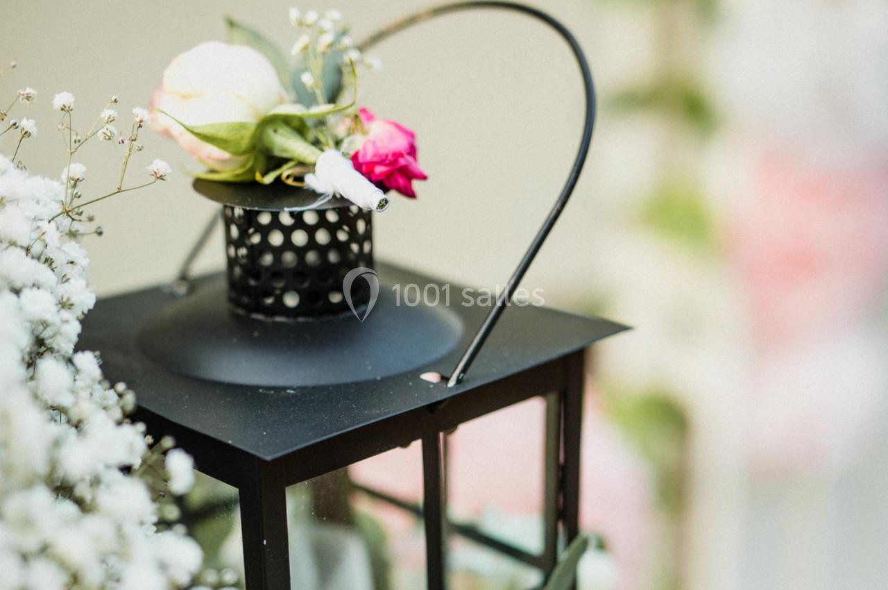 Lanterne noire décorée de petites fleurs blanches, roses et feuillage, posée dans un cadre lumineux et floral.