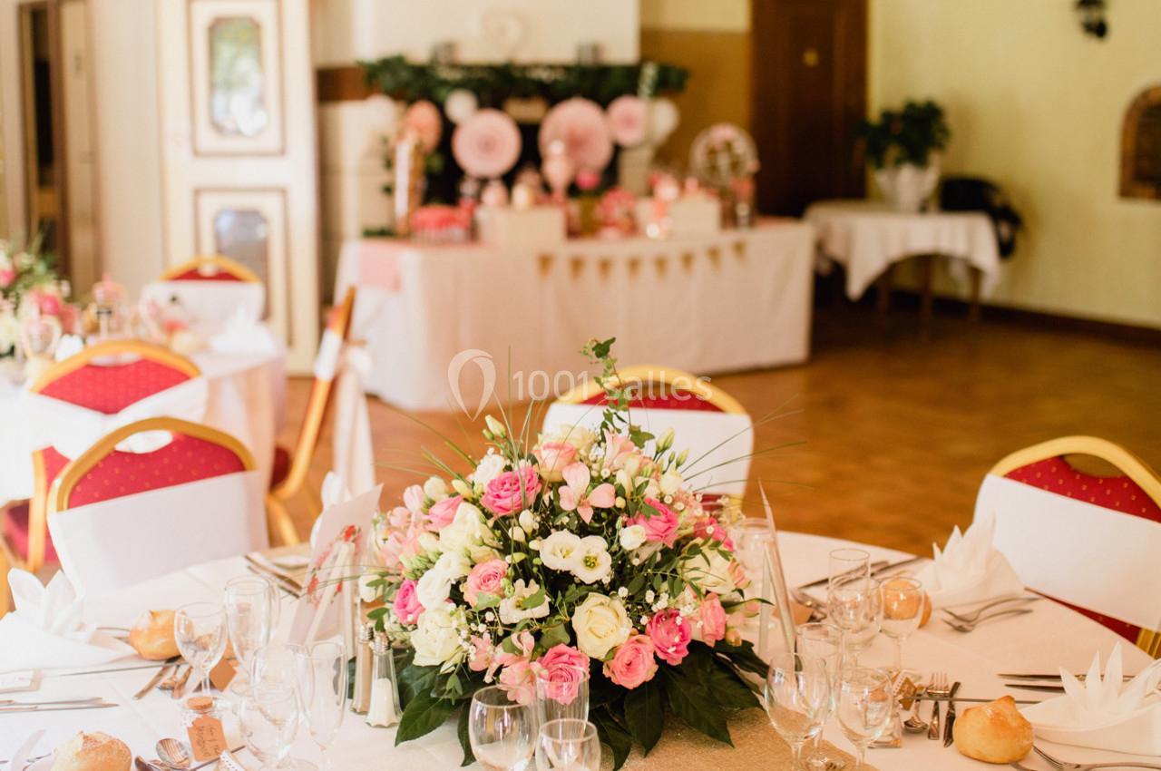 Salle de réception décorée pour un événement, avec des tables dressées et un centre de table floral rose et blanc.