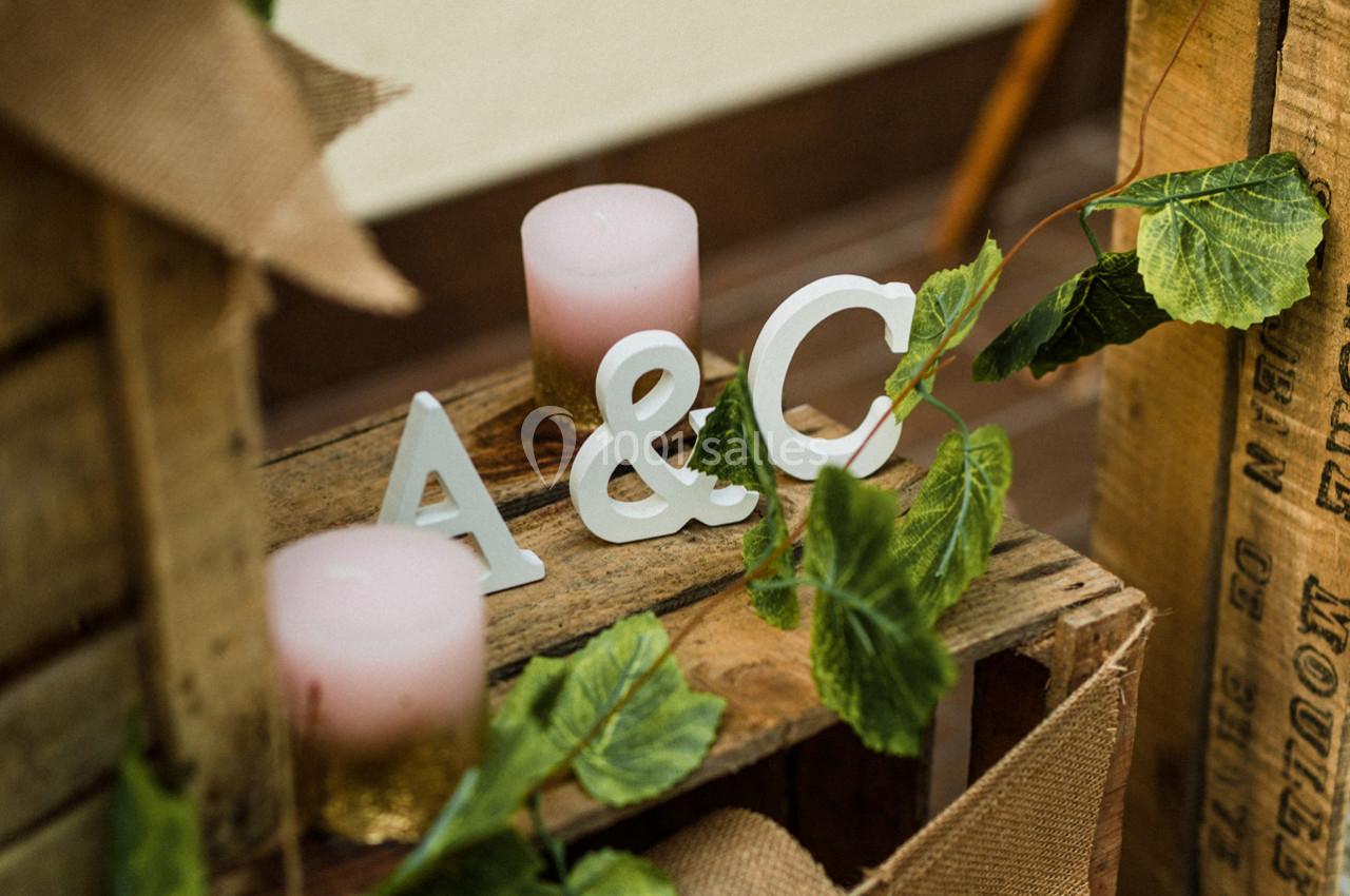 Bougies roses, lettres ’A & C’ blanches et feuillage décoratif posés sur des caisses en bois.