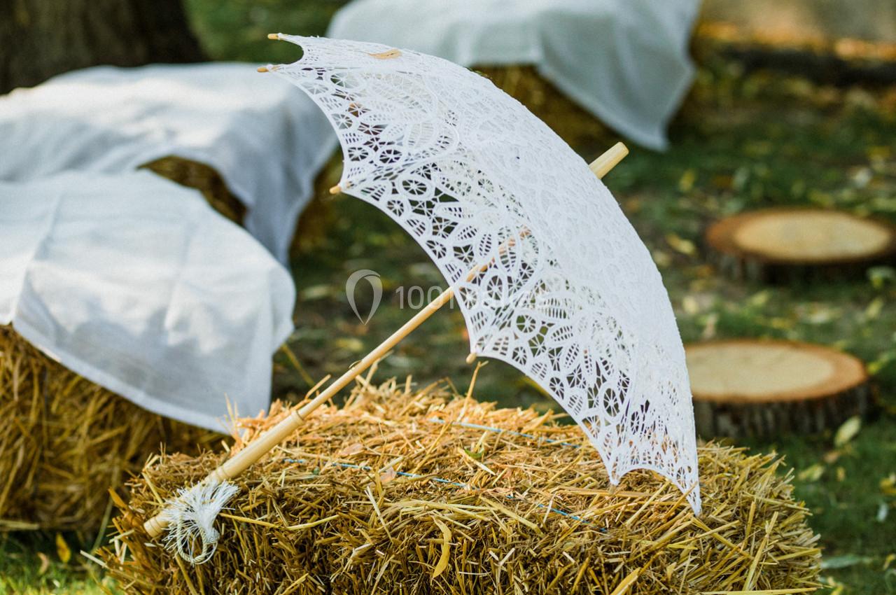Ombrelle blanche en dentelle posée sur une botte de paille dans un décor extérieur champêtre.