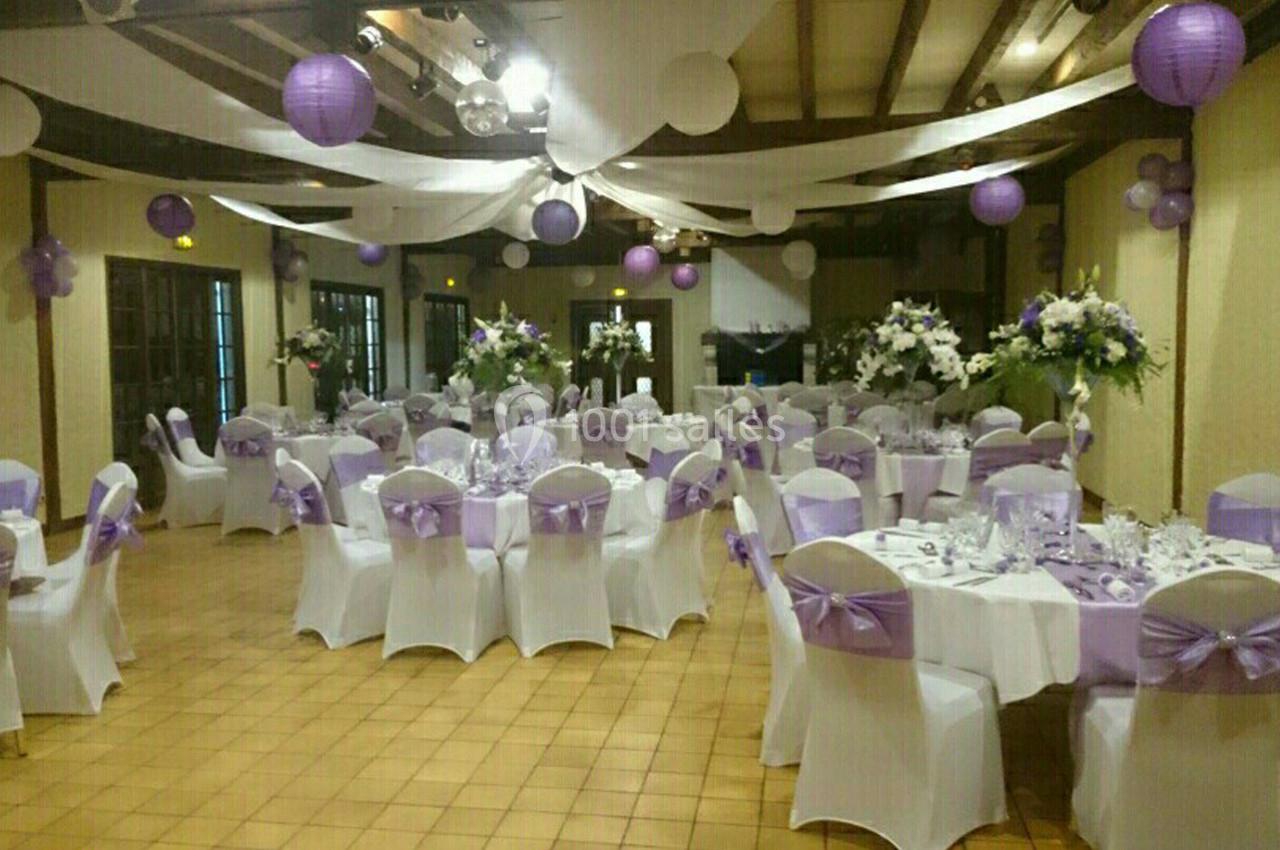Salle de réception décorée avec des tables rondes, nappes blanches, chaises ornées de rubans violets et lanternes suspendues.