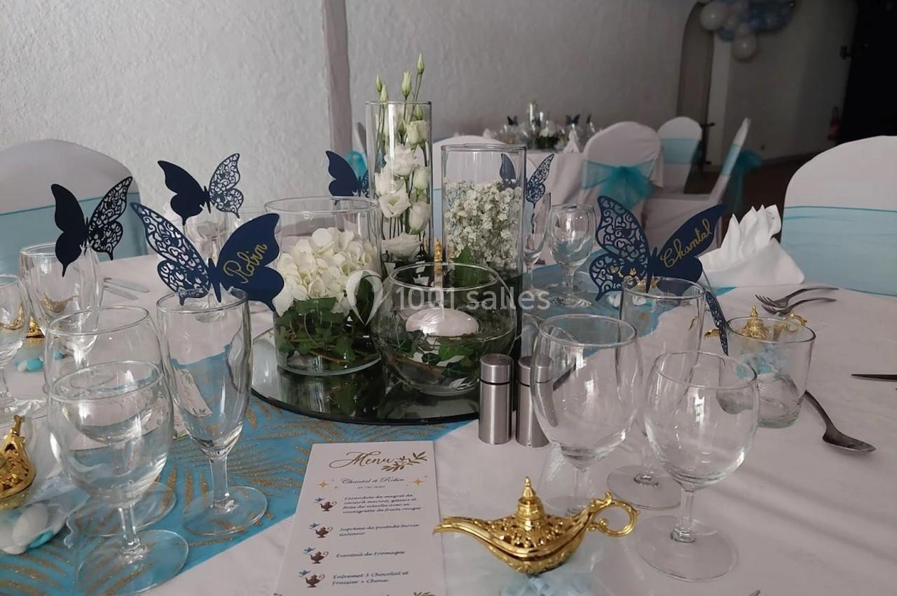 Table décorée pour un événement avec des fleurs blanches, papillons en papier bleu, lampes dorées et menu imprimé.