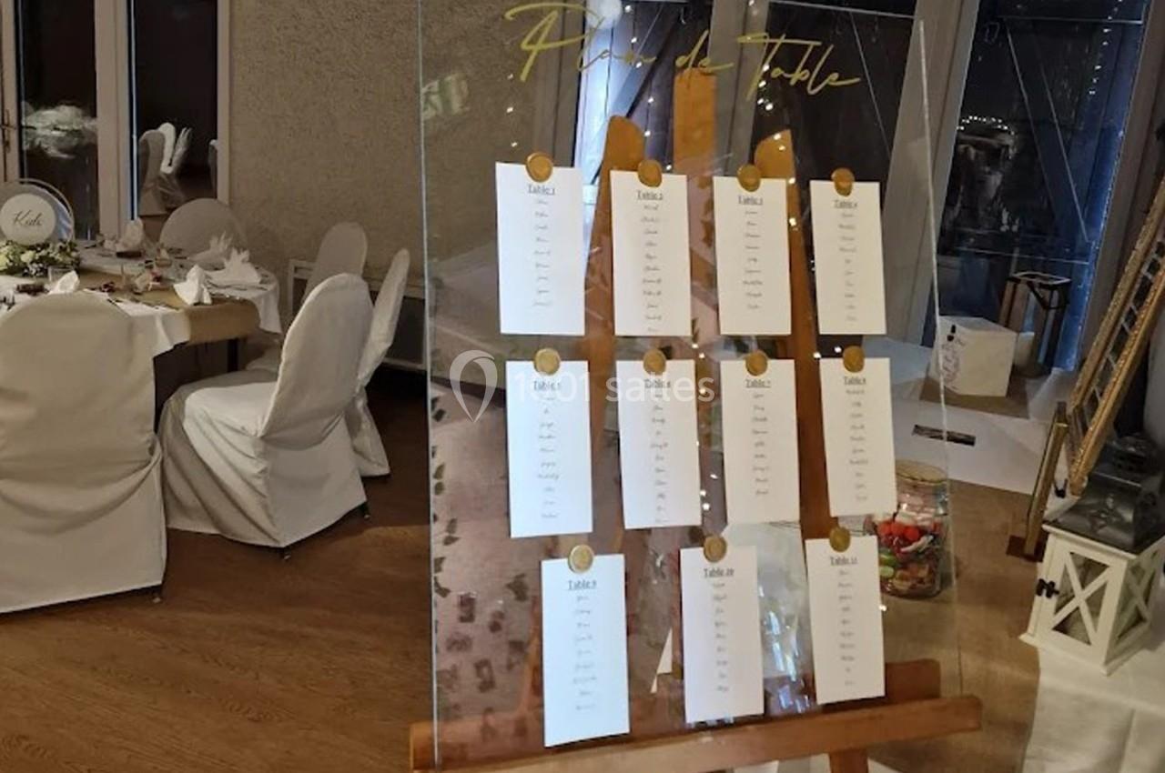 Plan de table sur un panneau transparent avec des cartes suspendues, placé sur un chevalet dans une salle de réception.