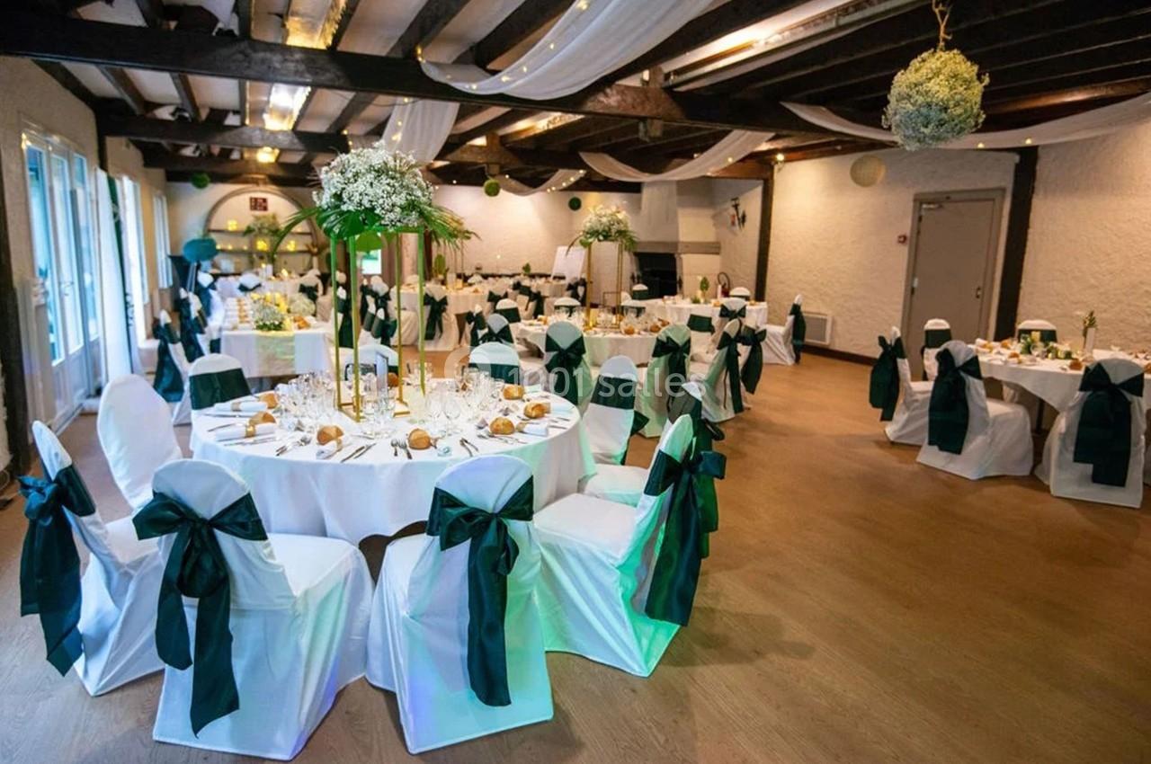 Salle de réception décorée avec des tables rondes, nappes blanches, chaises ornées de nœuds verts et éclairage chaleureux.